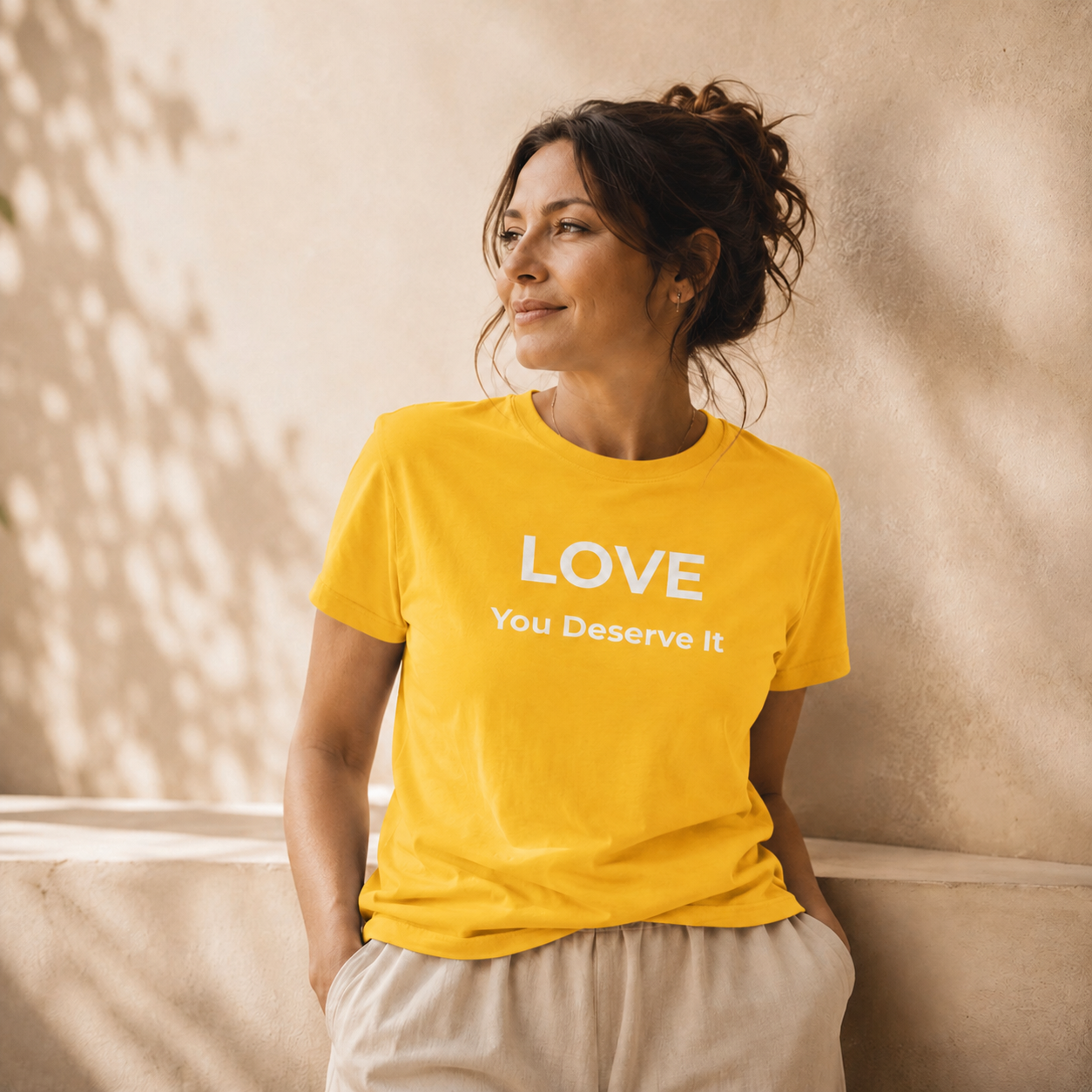 Woman wearing a yellow t-shirt with 'LOVE You Deserve It' text against a beige wall.