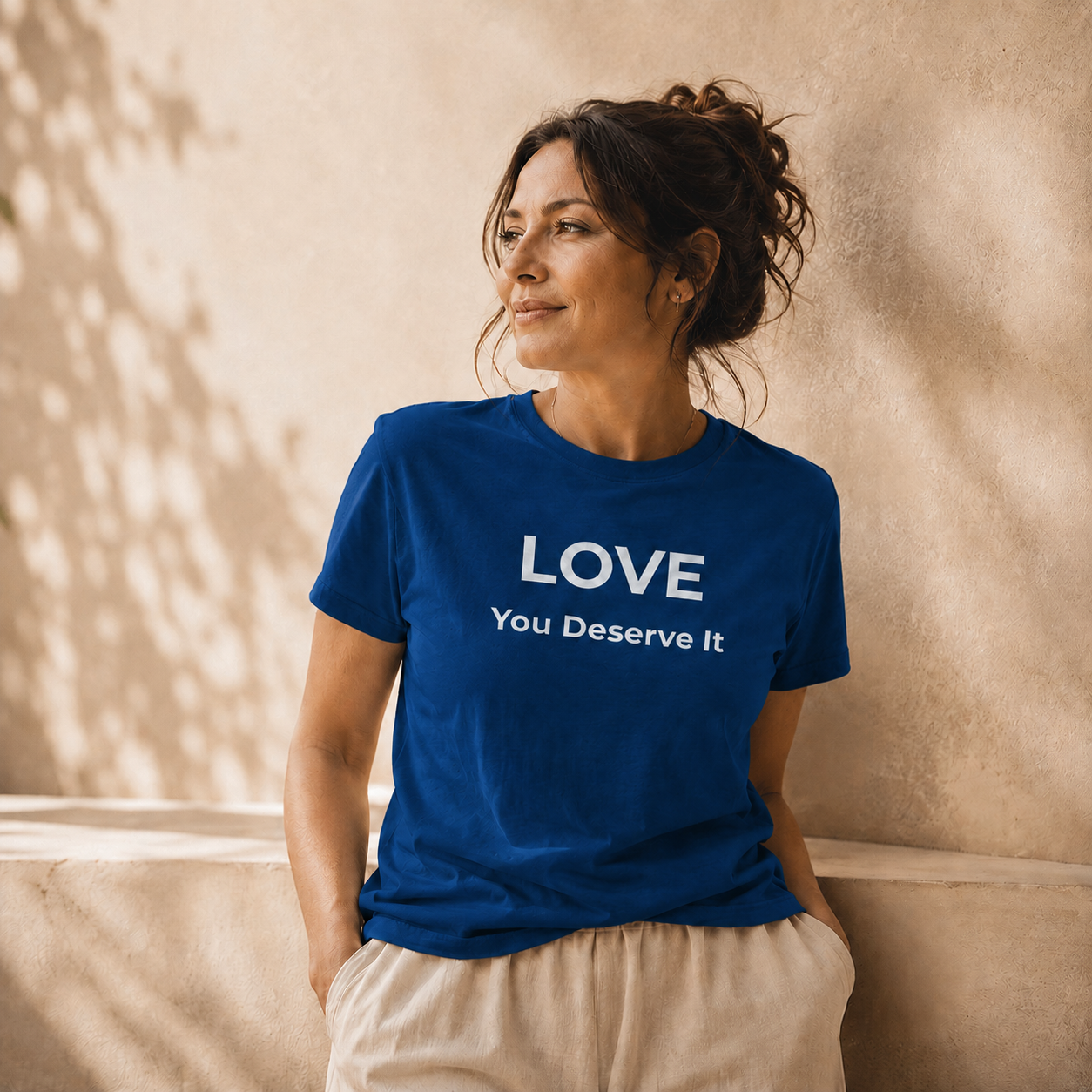 Woman wearing a blue t-shirt with 'LOVE You Deserve It' text against a beige wall.