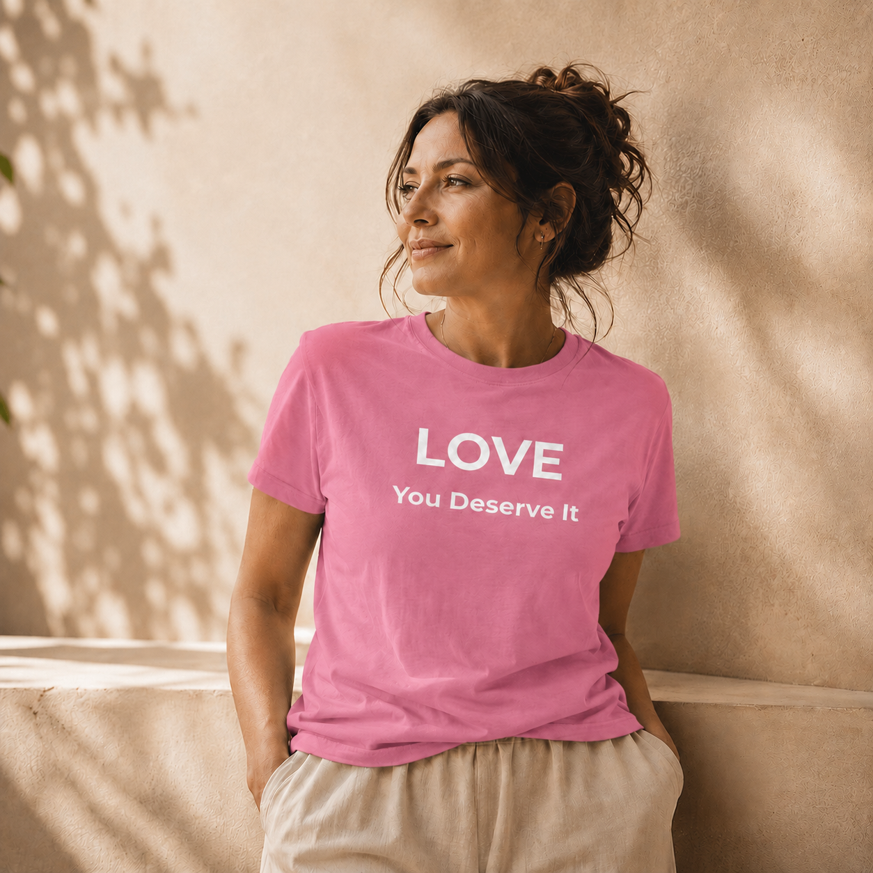 Woman wearing a pink t-shirt with 'LOVE You Deserve It' text against a beige wall.
