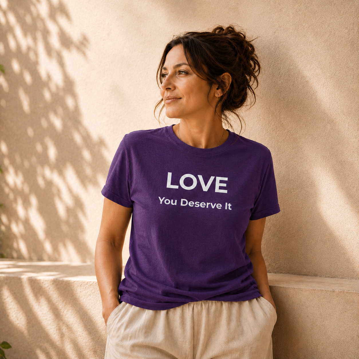 Woman wearing a purple t-shirt with 'LOVE You Deserve It' text against a beige wall.
