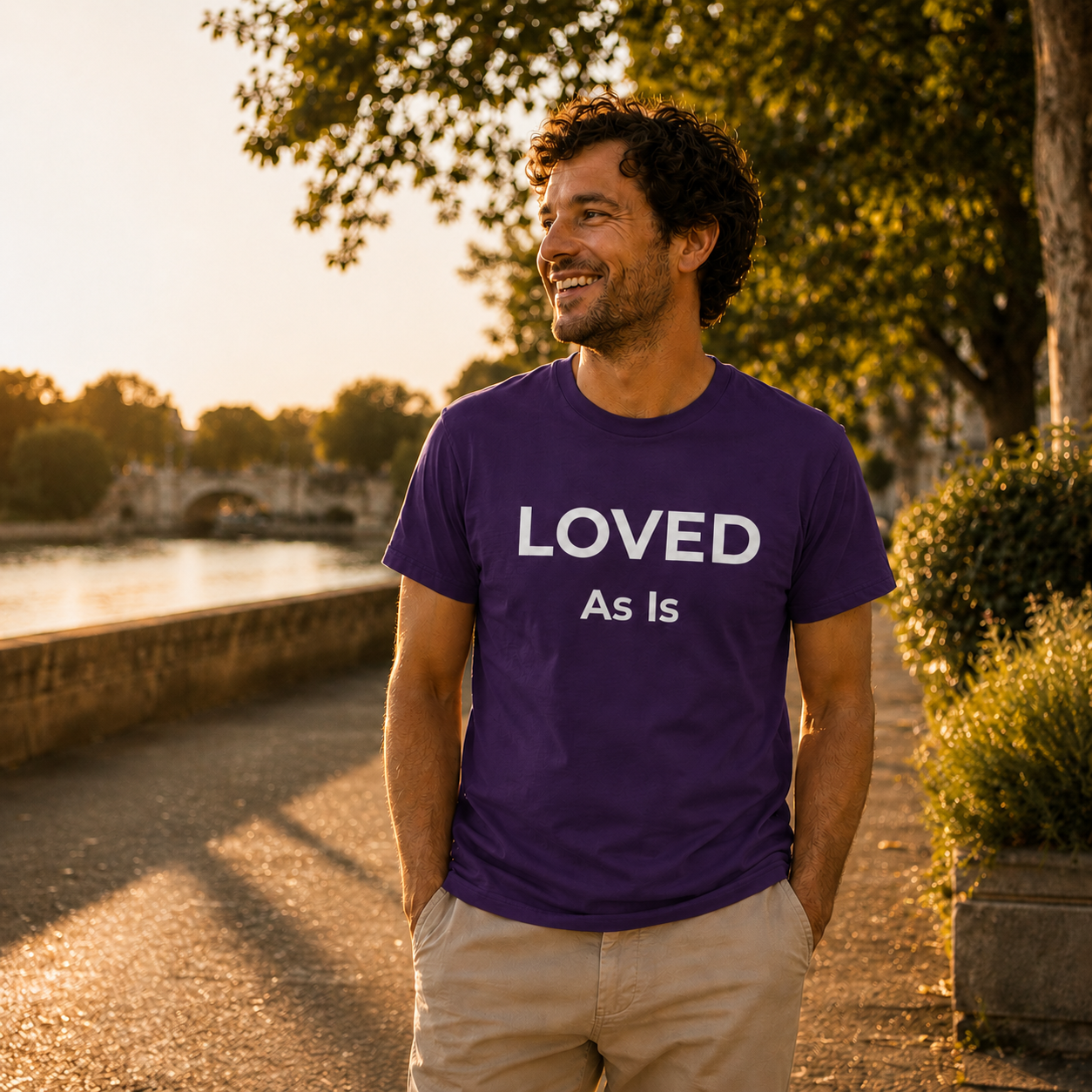 Man wearing a purple t-shirt with 'LOVED As Is' text, standing outdoors near a body of water.