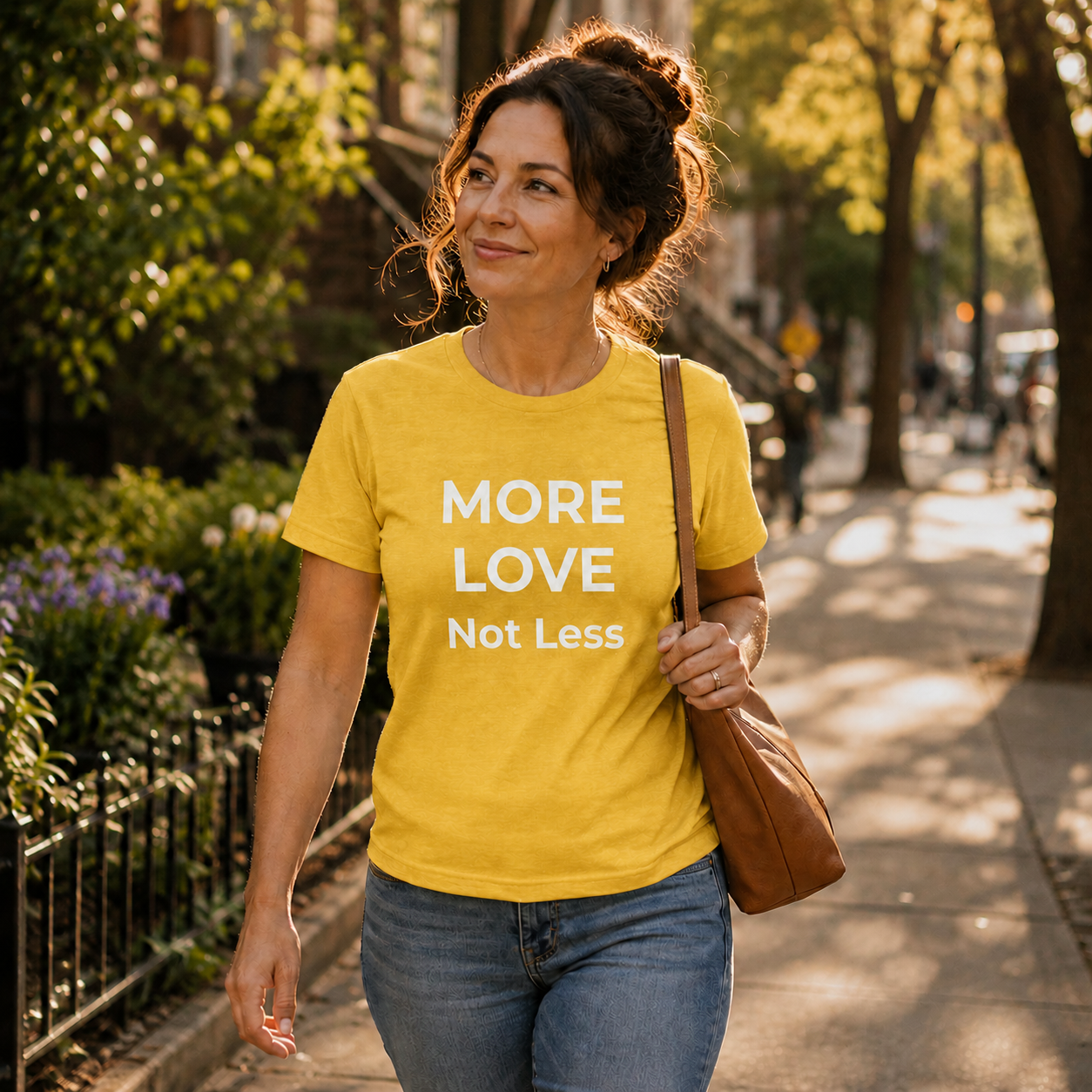 Woman walking on a sidewalk wearing a yellow t-shirt with 'MORE LOVE Not Less' text.