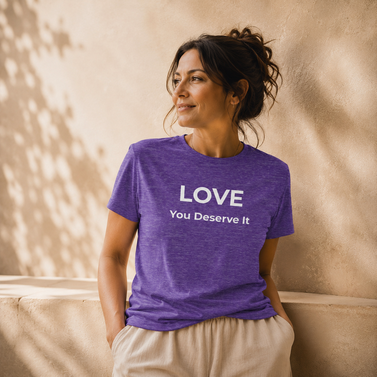 Woman wearing a heather purple t-shirt with 'LOVE You Deserve It' text against a beige wall.