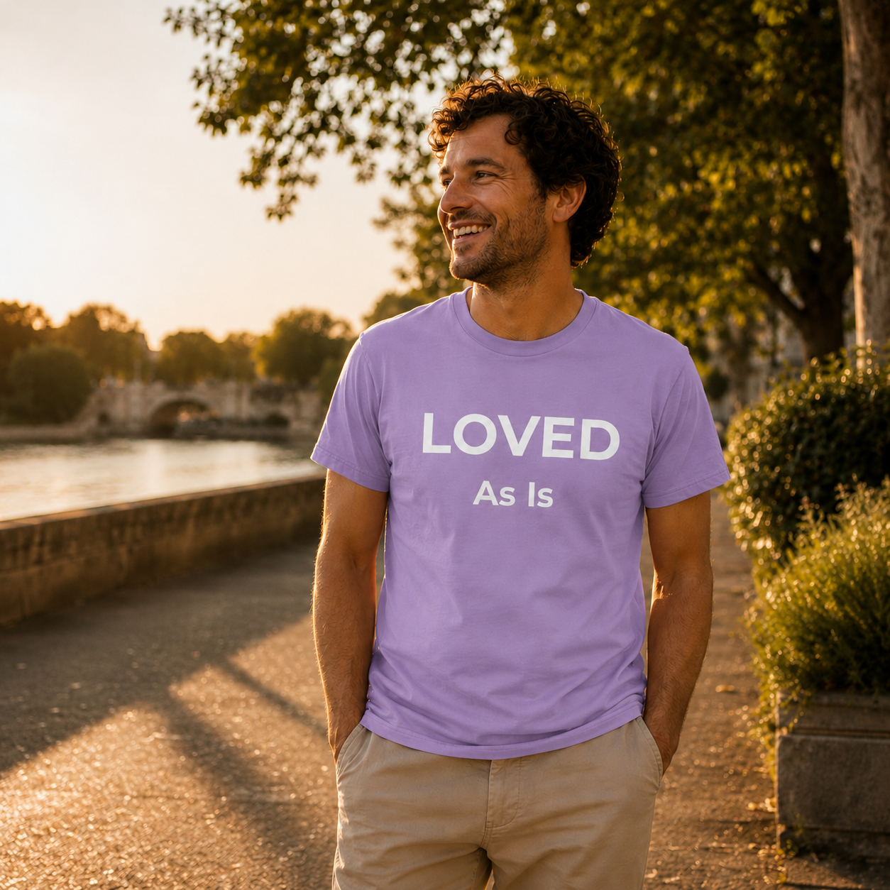 Man wearing a light purple t-shirt with 'LOVED As Is' text, standing outdoors near a body of water.