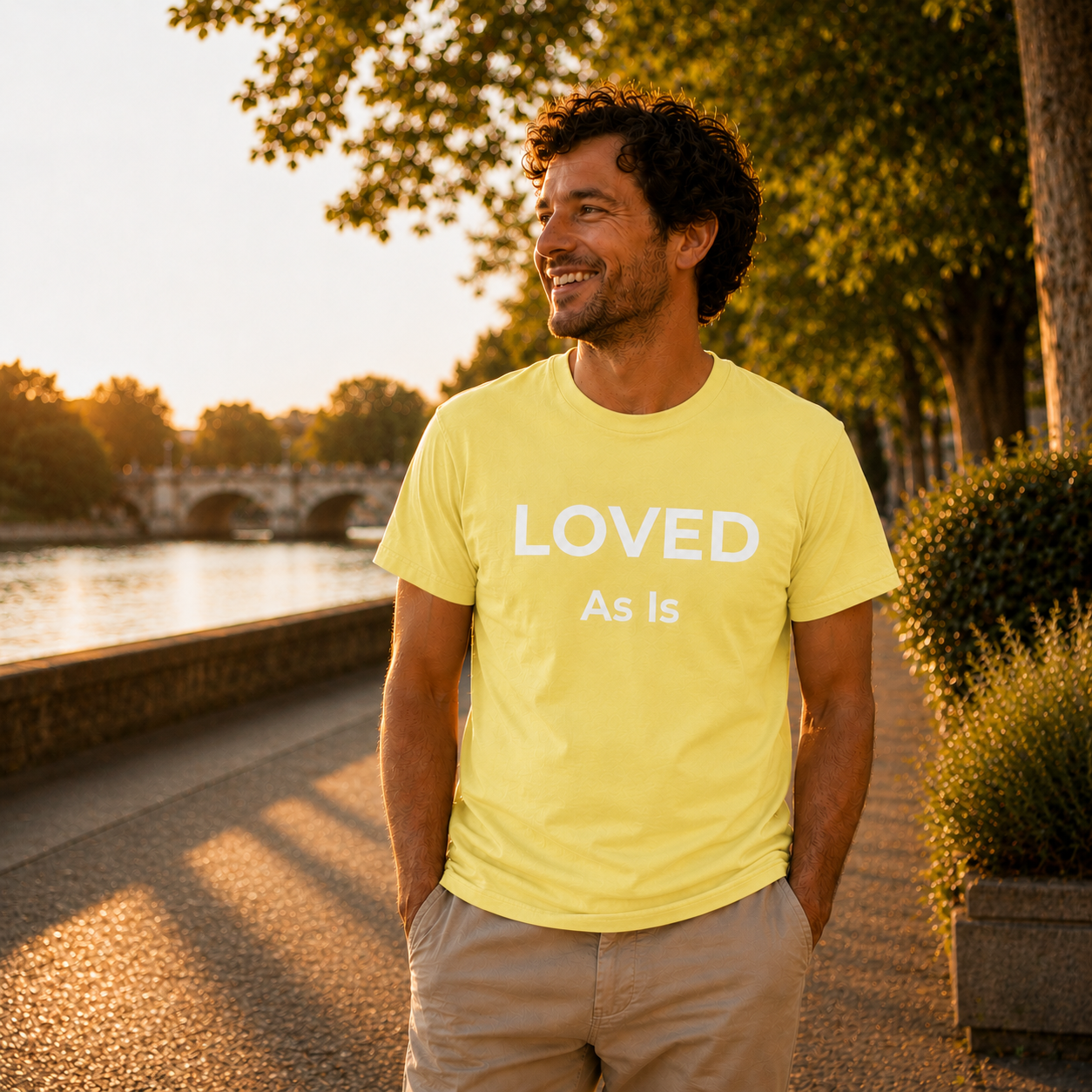 Man wearing a cornsilk yellow t-shirt with 'LOVED As Is' text, standing outdoors by a river.