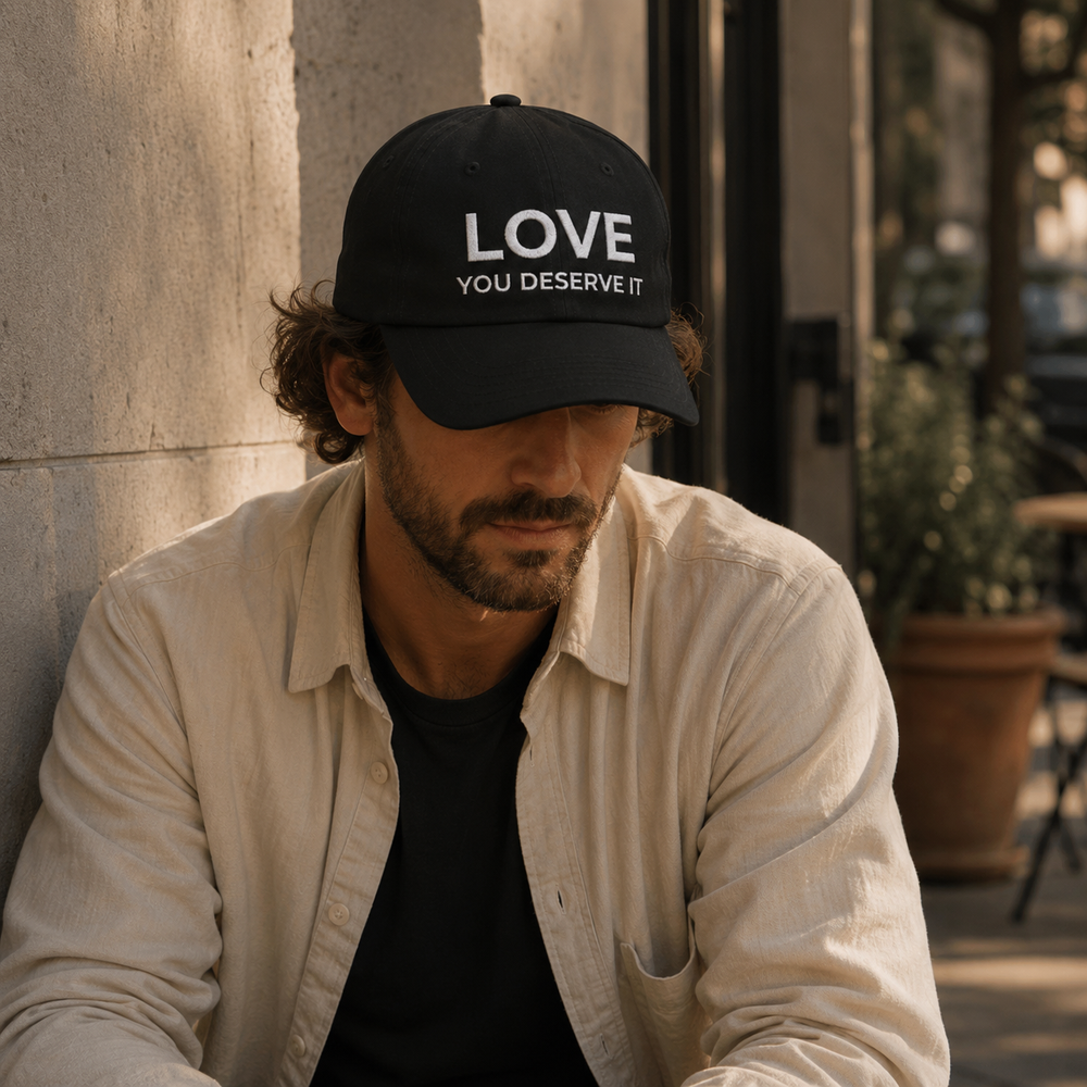 Man wearing a black cap with 'LOVE YOU DESERVE IT' text, sitting outdoors.