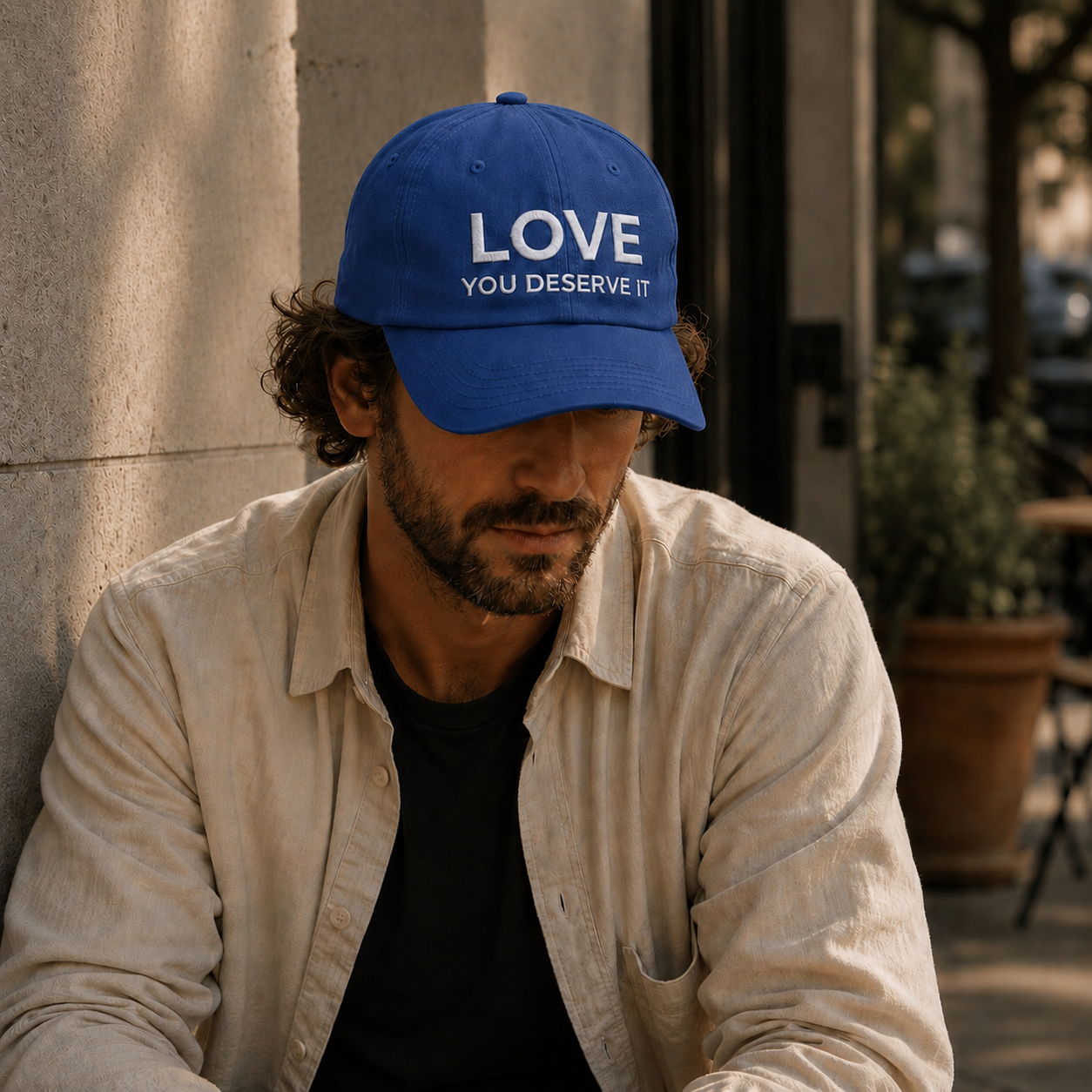 Man wearing a blue cap with 'LOVE YOU DESERVE IT' text, sitting outdoors.