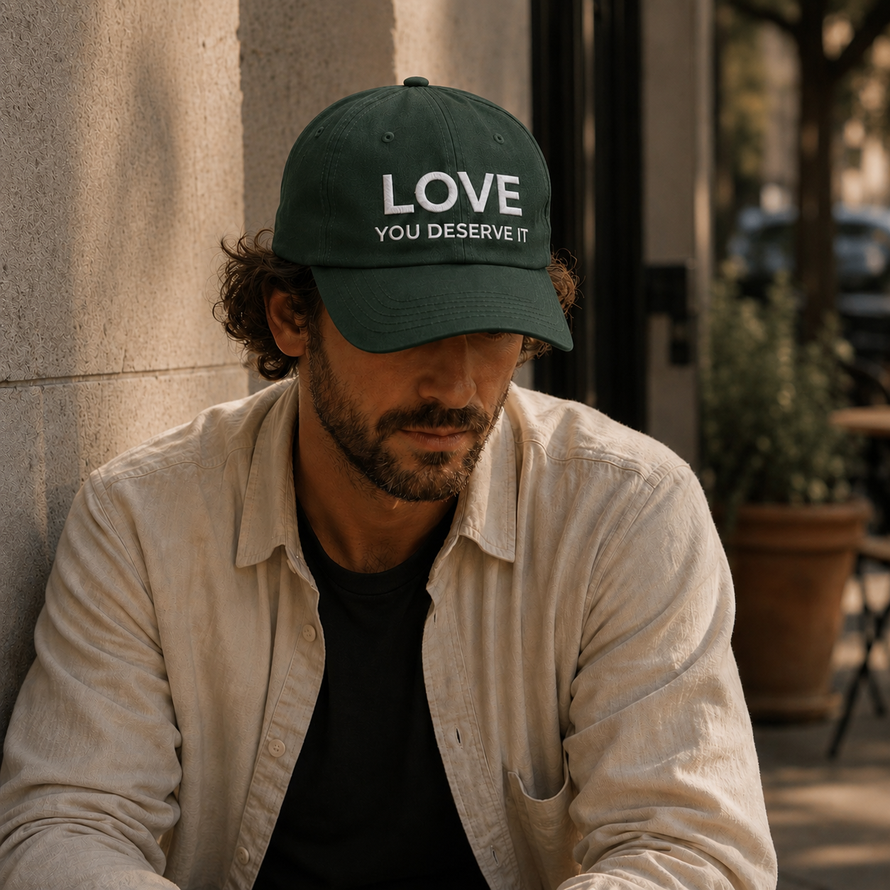 Man wearing a dark green cap with 'LOVE YOU DESERVE IT' text, sitting against a wall.