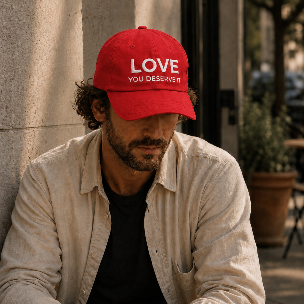 Man wearing a red cap with 'LOVE YOU DESERVE IT' text, sitting outdoors.