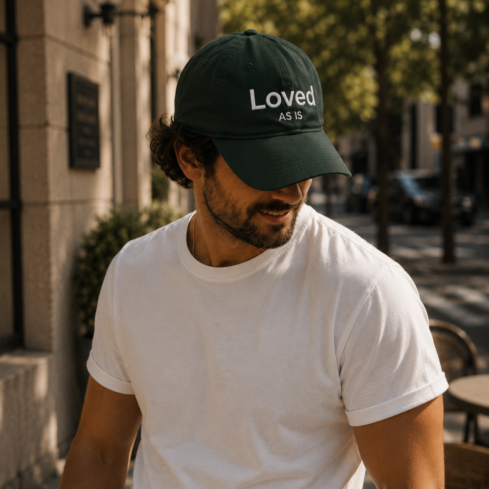 Man wearing a dark green cap with 'Loved As Is' text, standing outdoors.