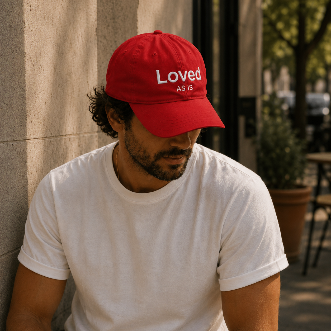 Man wearing a red cap with 'Loved As Is' text, sitting against a wall.