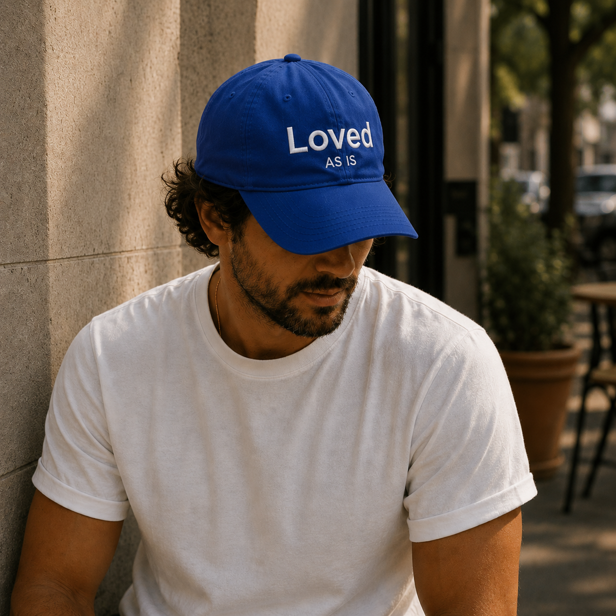 Man wearing a blue cap with 'Loved AS IS' text, sitting against a wall.