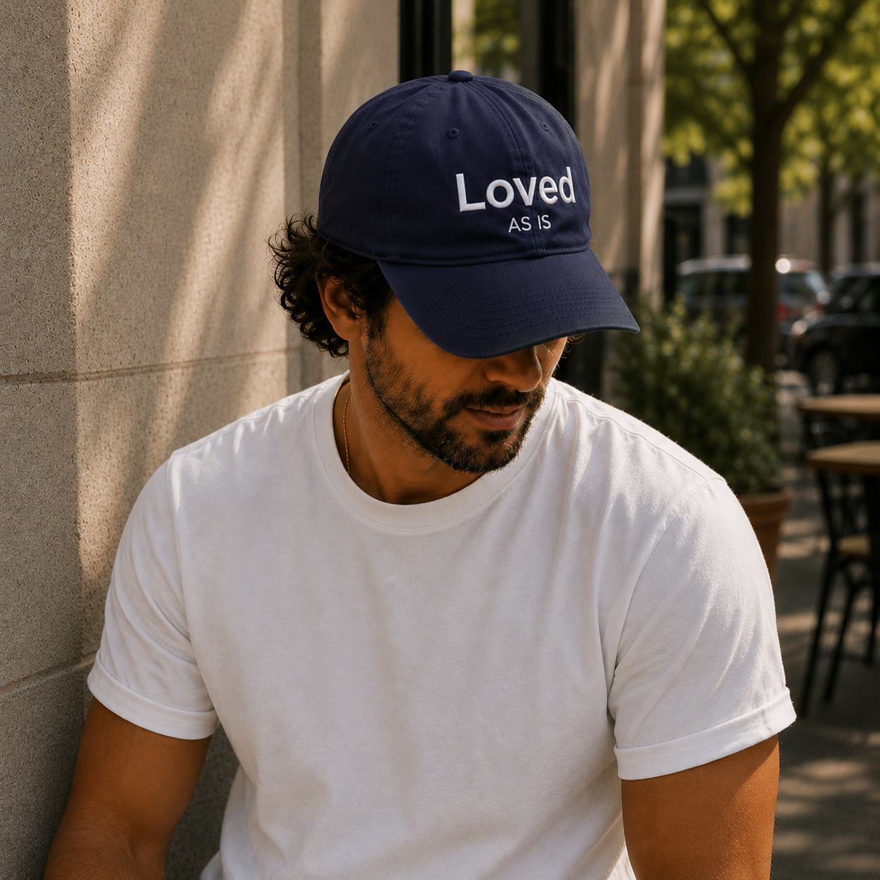Man wearing a navy blue cap with 'Loved As Is' text, sitting outdoors.