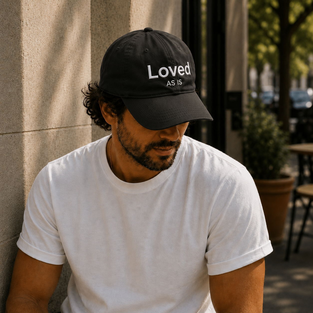 Man wearing a black cap with 'Loved As Is' text, sitting against a wall.